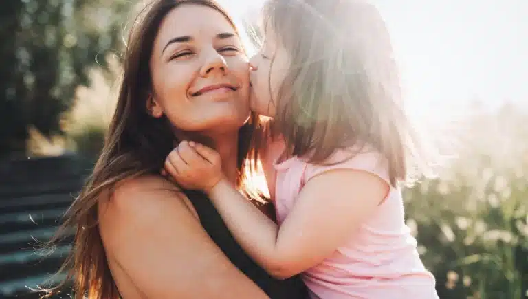 Brünette lächelnde Frau wird von einem kleinen Mädchen mit besonderen Bedürfnissen auf die Wange geküsst