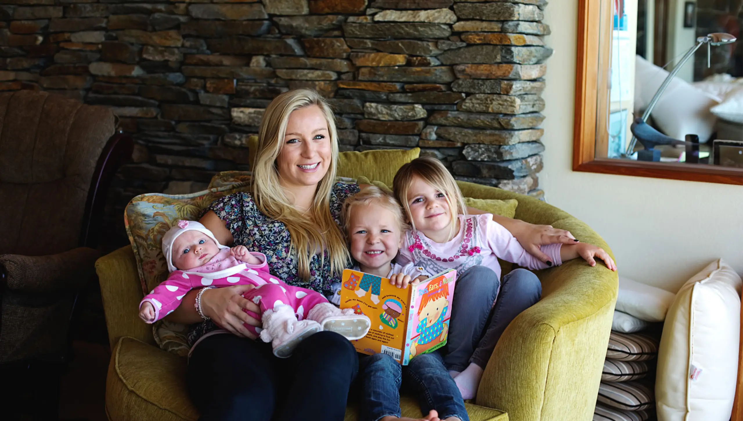 Junge blonde Frau sitzt auf einem Sofastuhl und hält ein Baby und zwei Kleinkinder